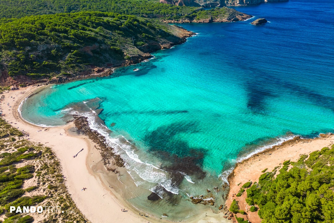 Aerial shot of Cala Algaiarens with shallow turquoise waters and sandy beach in Menorca, Spain