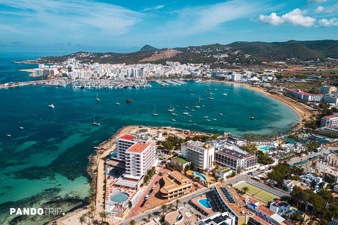 Aerial shot of San Antonio harbor in Ibiza with boats, beachfront hotels, and deep-blue Mediterranean sea.