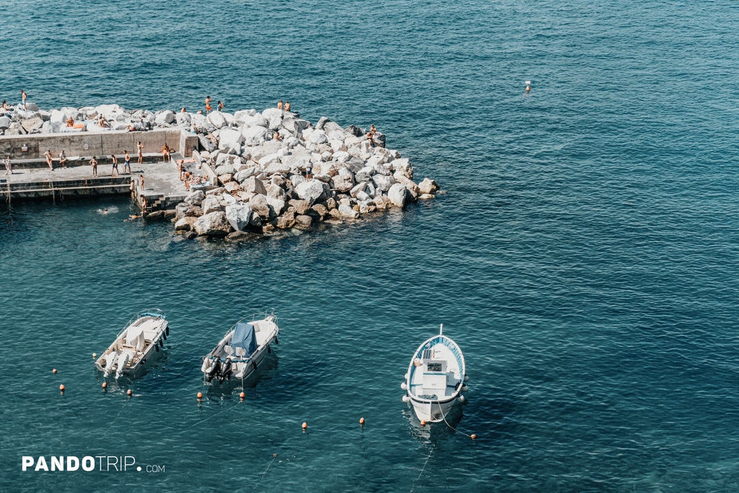 Small boats floating in the clear blue harbor of Vernazza with people sunbathing on the rocks