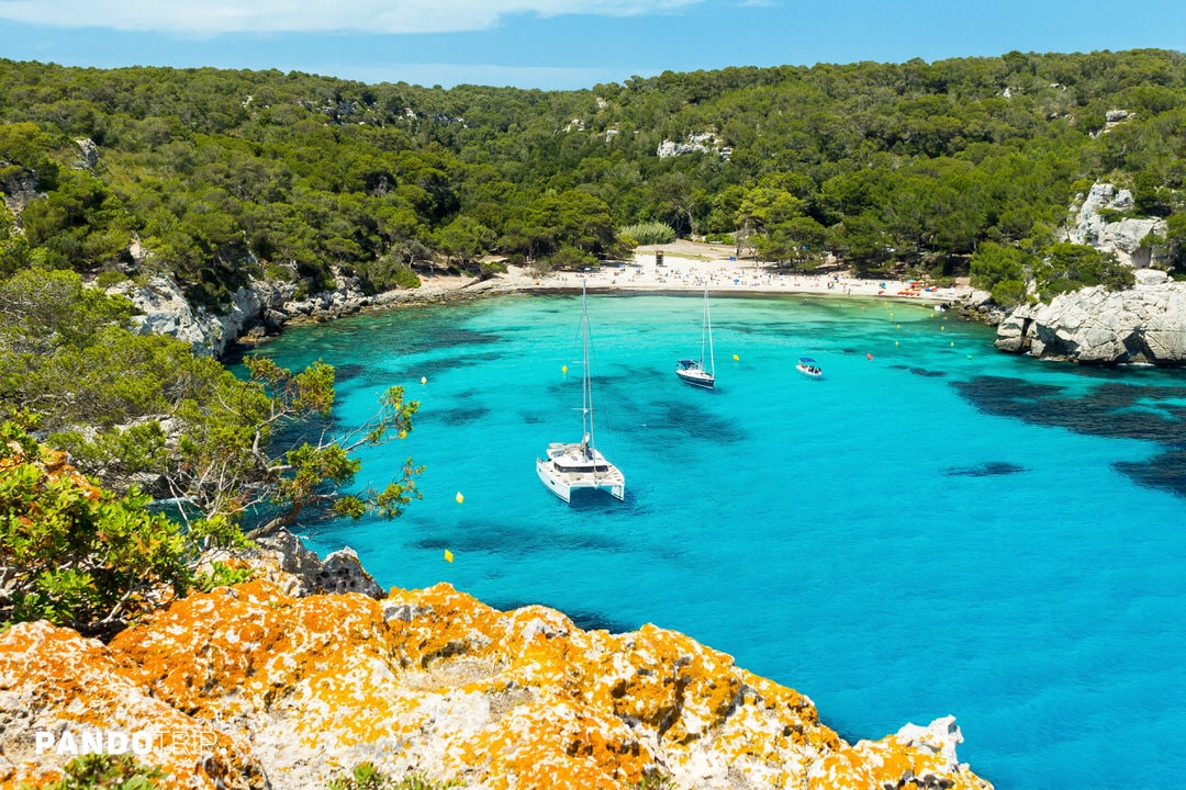View of Cala Macarella beach with turquoise waters and anchored boats in Menorca, Spain
