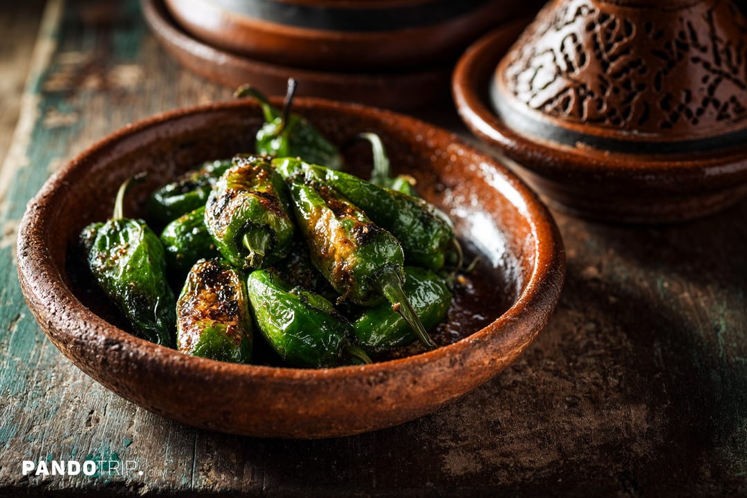 A small rustic bowl piled with blistered green padrón peppers, sea salt crystals visible on the skins
