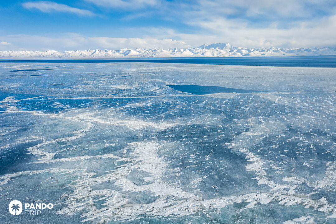 Aerial view of the turquoise frozen edge of Namtso Lake meeting the rocky desert-like Tibetan plateau.