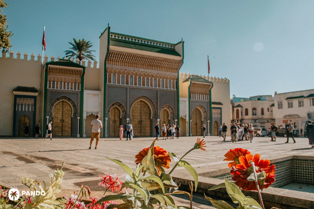 Tourists visiting the ornate golden gates of the Royal Palace in Fes el-Bali with red flowers in the foreground
