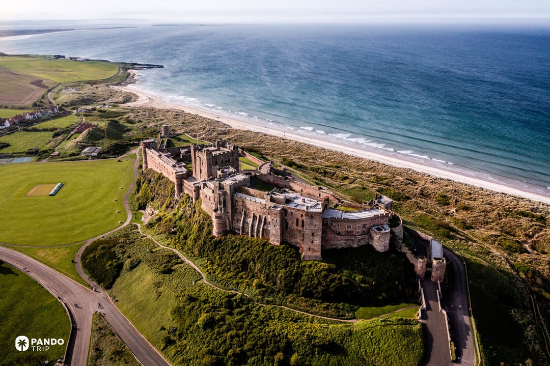 High-angle view of Bamburgh Castle on a green headland above the North Sea beach.