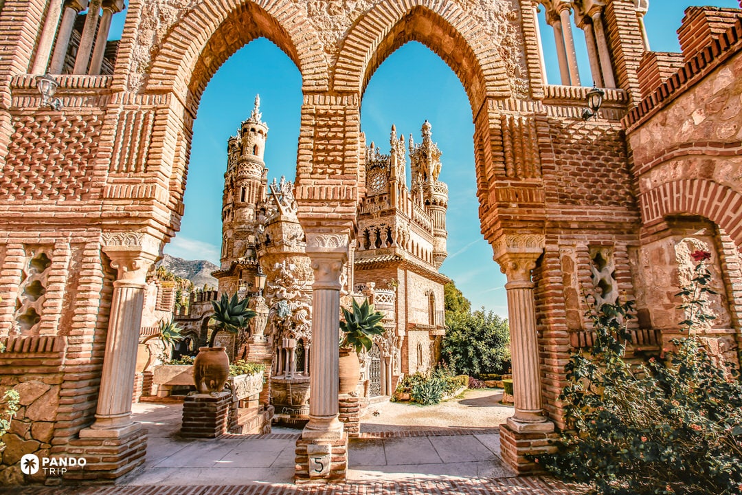 Brick arch frames the eclectic towers of Castillo de Colomares under blue sky.