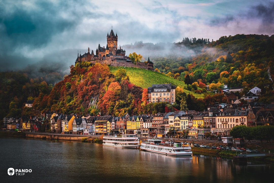 Cochem Castle rising above autumn vineyards and the Moselle River with colourful town below.