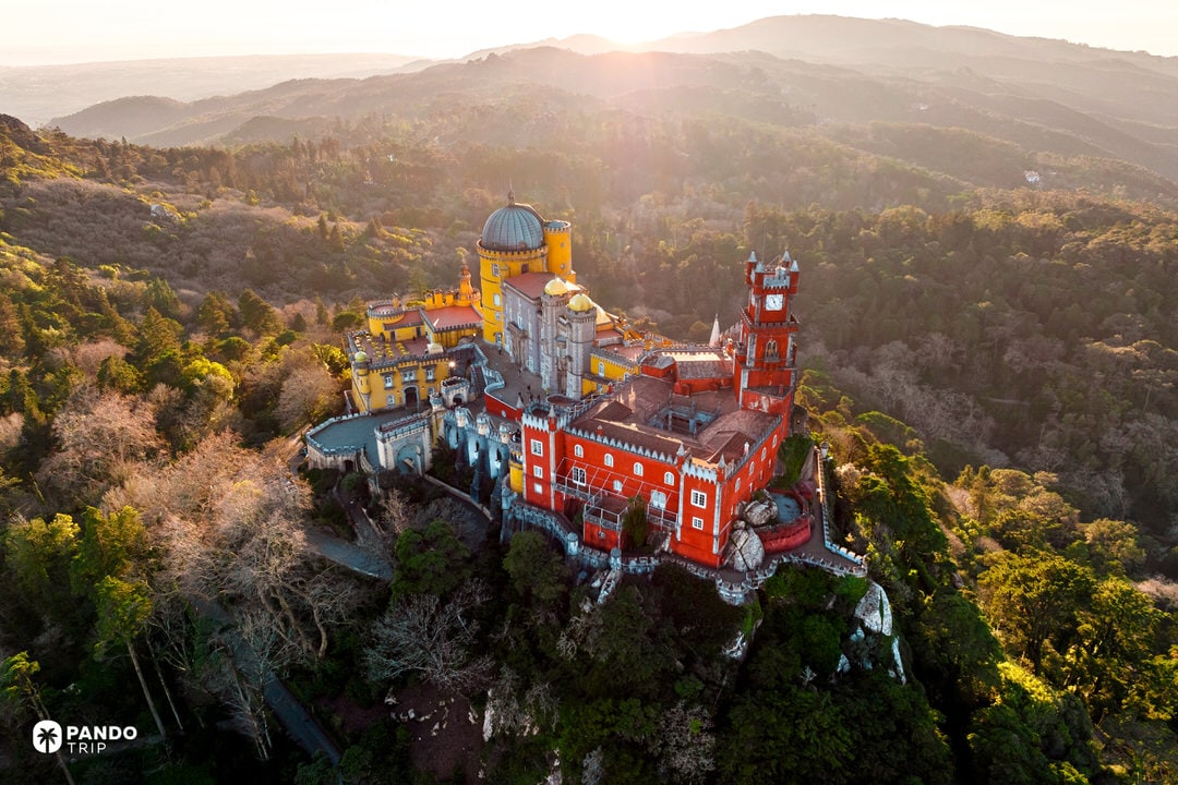 Sunset aerial of Pena Palace’s red and yellow towers rising from Sintra’s forested ridge.