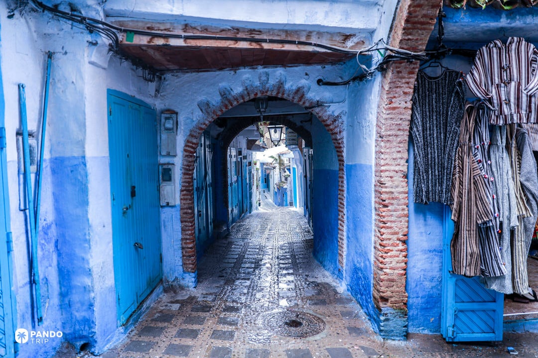 Narrow blue-painted alley with arched doorway in Chefchaouen’s medina, lined with shops and traditional doors