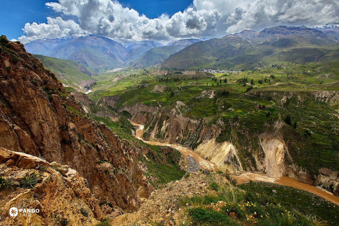 Green agricultural terraces stepping down toward the Colca Canyon, Peru, with villages and ravines beneath growing afternoon clouds.