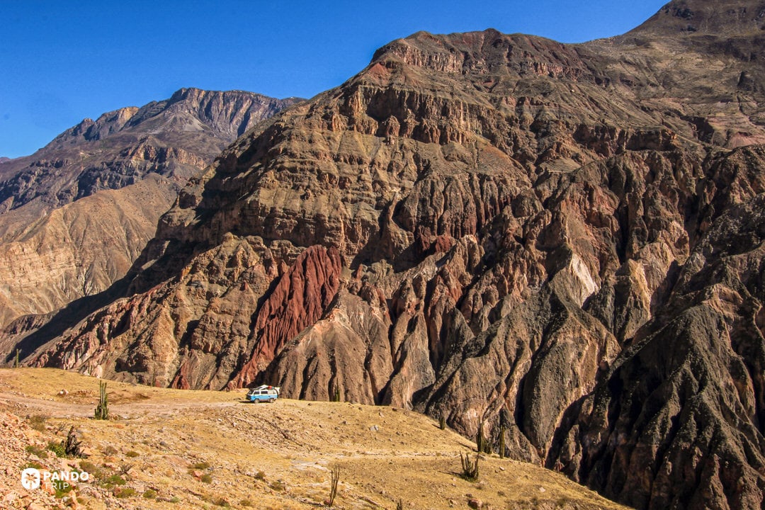 Layered volcanic cliffs of Cotahuasi Canyon in Peru under a clear Andean sky, tiny vehicle on the rim showing the canyon’s scale.