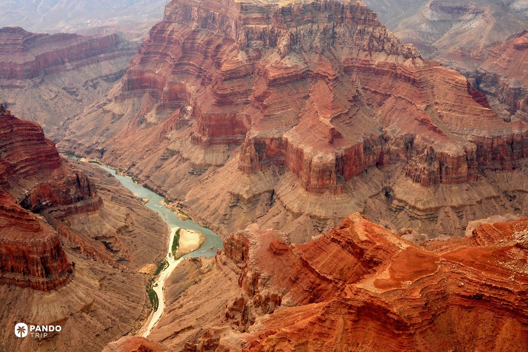 Colorado River winding below layered red cliffs in Grand Canyon National Park, Arizona, with a sandy river bend under hazy afternoon light.