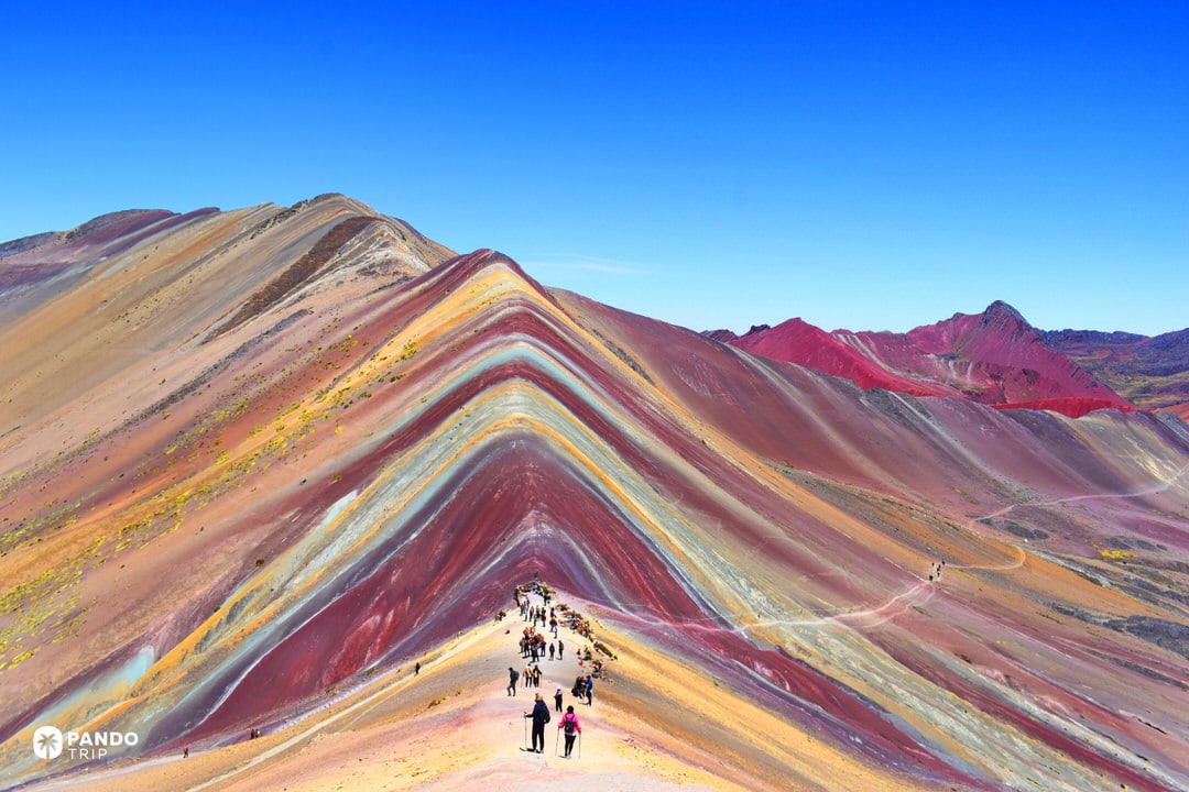 Hikers walking along the colorful ridge of Vinicunca (Rainbow Mountain) under a deep blue sky in Peru.
